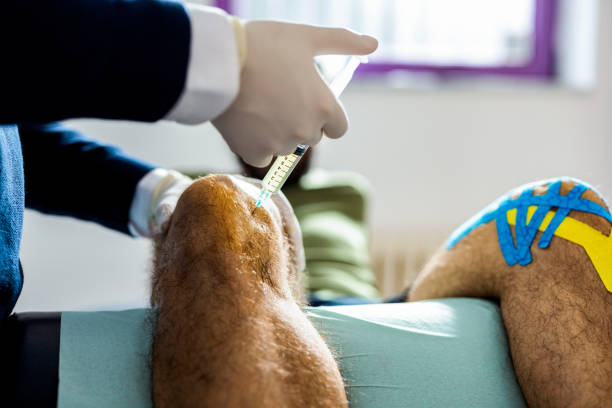 Close up of the patient's knee, doctor is injecting a fluid to the knee with a syringe and a needle. Unrecognizable doctor giving knee injection at his office while patient sitting at medical bed.