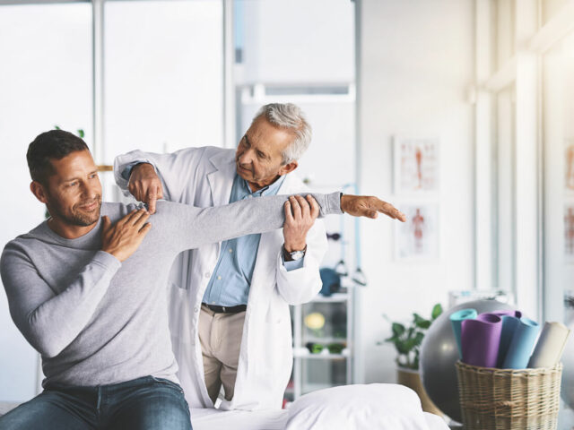 Male patient with doctor at orthopedic clinic performing exam on shoulder.