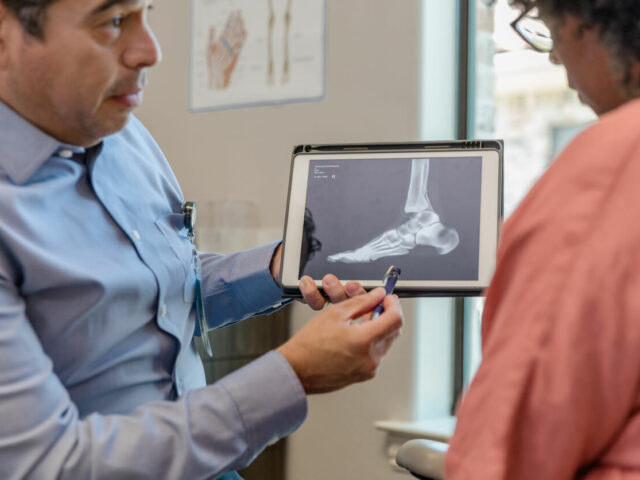 A senior orthopedic surgeon holding a tablet displaying a foot x-ray while listening to an elderly woman during orthopedic care.